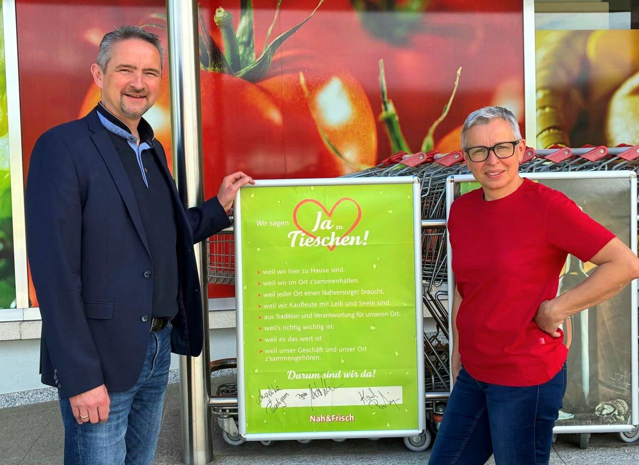 Zwei Männer stehen lächelnd neben einem grünen Schild. Das Schild trägt den Text 'Ja zu Tischen!' mit Gründen für die Unterstützung. Dahinter sind Einkaufswagen und eine große Tomaten-Display zu sehen.