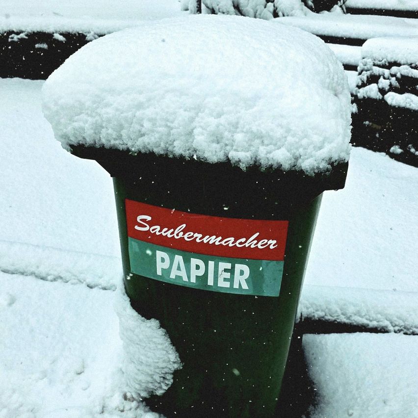 A green waste bin with a red label, covered in snow, is placed on a snowy surface. The label reads Saubermacher Papier.