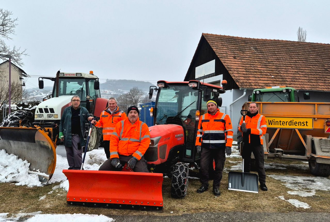 Five men in orange safety jackets and gloves pose next to machinery vehicles. One man holds a snowplow, another a shovel. Behind them, a building with a brown roof and a tractor.