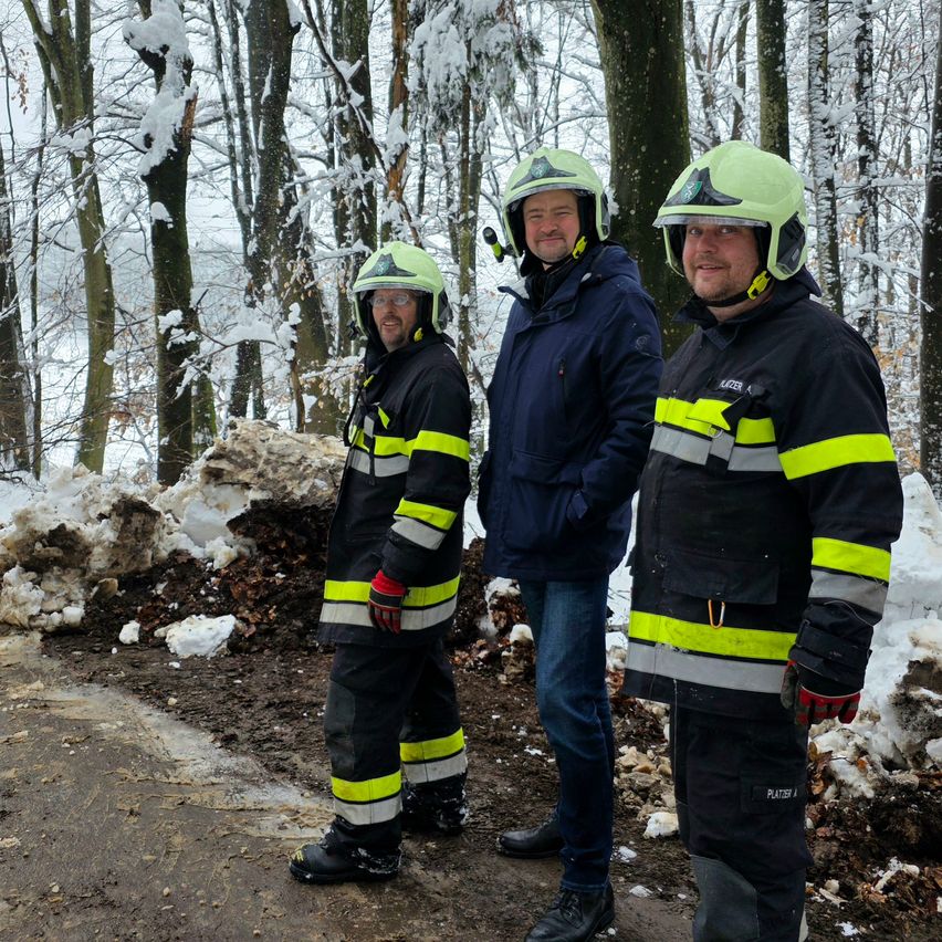 Drei Männer stehen auf einem verschneiten Pfad in einem Wald. Sie tragen Sicherheitsanzüge und Helme, einer hält eine Kamera. Mit schneebedeckten Bäumen umgeben sie.