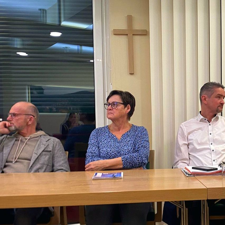 Drei Personen sitzen an einem Tisch in einem Besprechungsraum. Ein Mann trägt eine Brille und eine graue Kapuzenjacke. Eine Frau trägt eine Brille und eine blaue Bluse. Ein anderer Mann trägt ein weißes Hemd. Ein Kreuz hängt an der Wand.