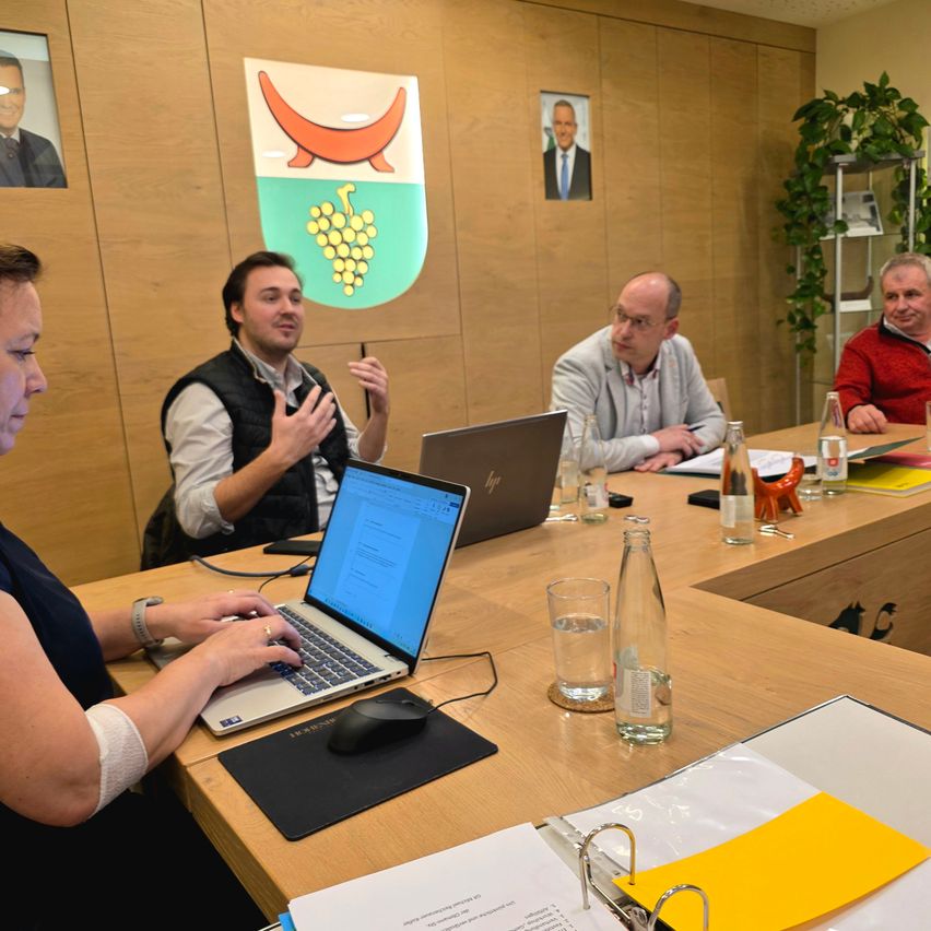 Eine Gruppe von Menschen sitzt in einem Besprechungsraum um einen Tisch herum. Laptops, Papiere und Flaschen sind auf dem Tisch. Ein Mann in der Mitte spricht und gestikuliert. Ein anderer Mann sitzt am Tisch und schaut den Mann an. Eine Frau sitzt links und tippt auf ihrem Laptop.