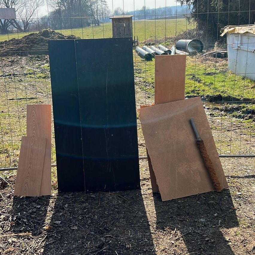 Mehrere Holzbretter sind neben einem Zaun gestapelt. Ein großes schwarzes Brett und einige braune Papiere sind auf dem Boden angeordnet. Hinter dem Zaun befindet sich ein kleines Haus und einige Rohre.