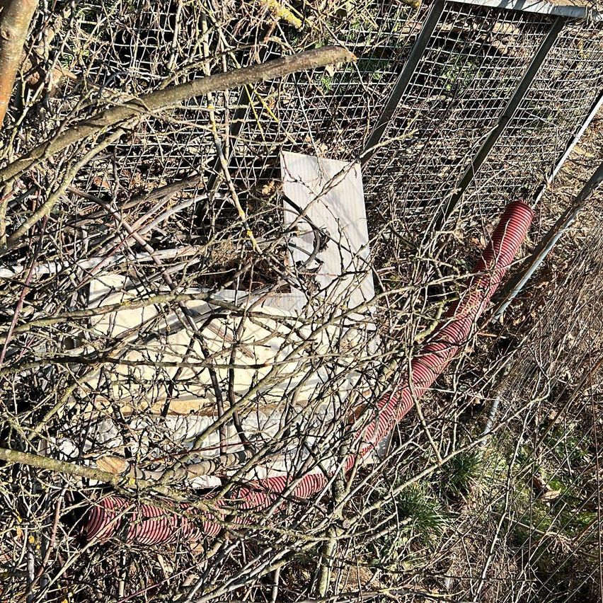 Ein rostiger Metallzaun teilweise von Ästen und Zweigen bedeckt. Ein roter Schlauch ist sichtbar, und es gibt Überreste von Karton oder Papier zwischen den Ästen.