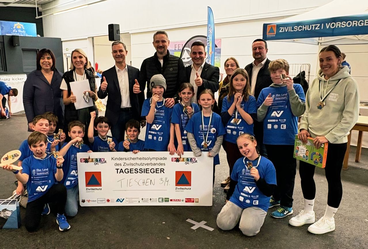 Eine Gruppe von Kindern und Erwachsenen posiert für ein Foto. Sie halten Medaillen und ein Banner in der Hand. Sie tragen passende blaue Shirts. Sie stehen in einem Raum mit einem blauen Banner im Hintergrund. Das Bild ist klar und gut beleuchtet.