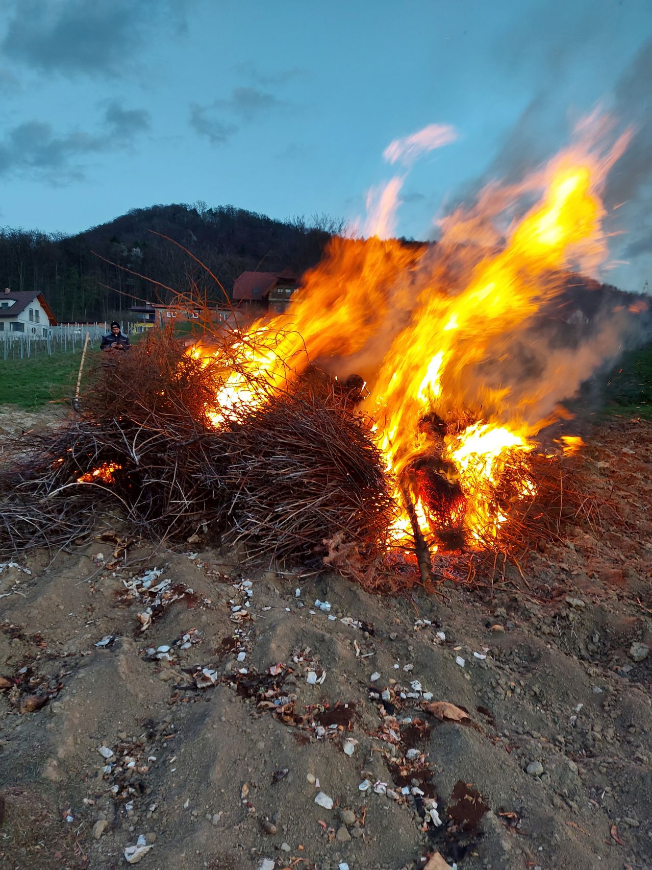 Ein großes Feuer brennt in der Mitte eines Haufens trockener Zweige und Äste auf einem Erdfeld. Im Hintergrund sind ein Berg und Häuser sichtbar.