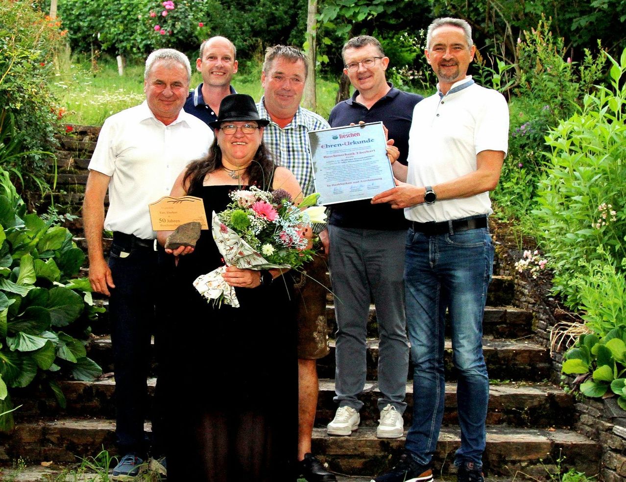 A group of people stands on a staircase, with a woman holding a bouquet and a plaque. The man on the right holds a certificate. They are all smiling and appear to be celebrating an event.