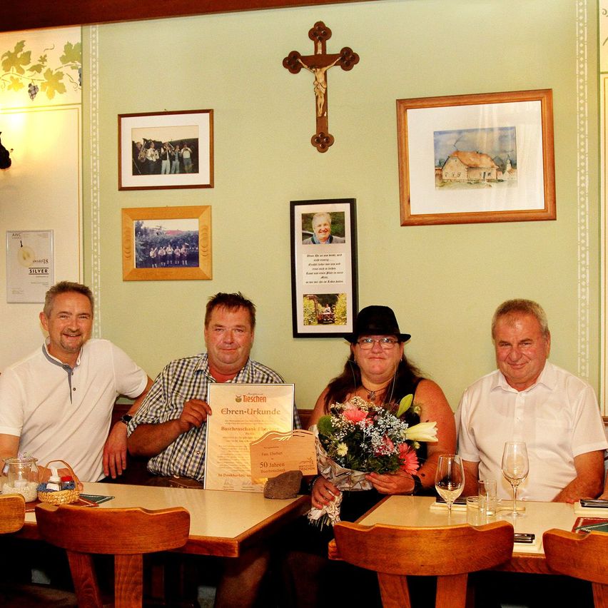 Four people are seated at a table in a restaurant, smiling for a photo. A woman holds a bouquet of flowers and a certificate. Behind them, a wall displays a cross and several framed pictures.