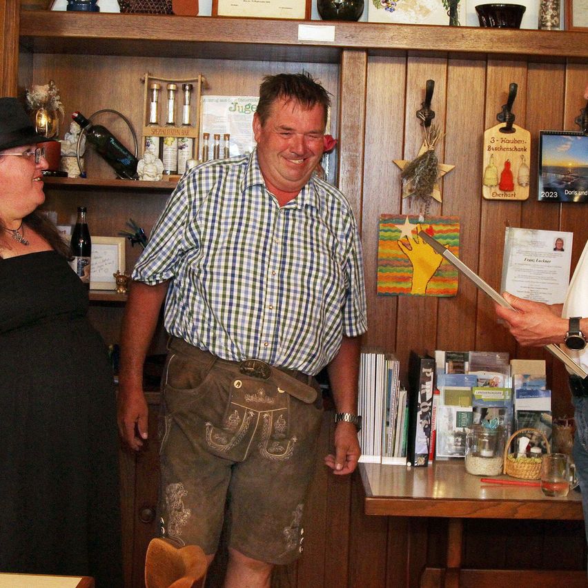 A man in traditional Bavarian clothing stands in a room with a woman and another man, smiling and looking at a paper. Behind him, a wooden shelf holds various items.