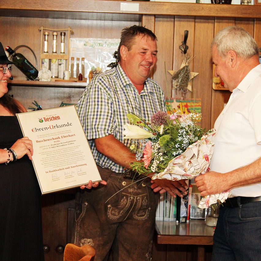 A man in traditional German attire holds a certificate and a bouquet of flowers while a woman and another man look on. The setting appears to be a room with wooden shelves and various items on display.