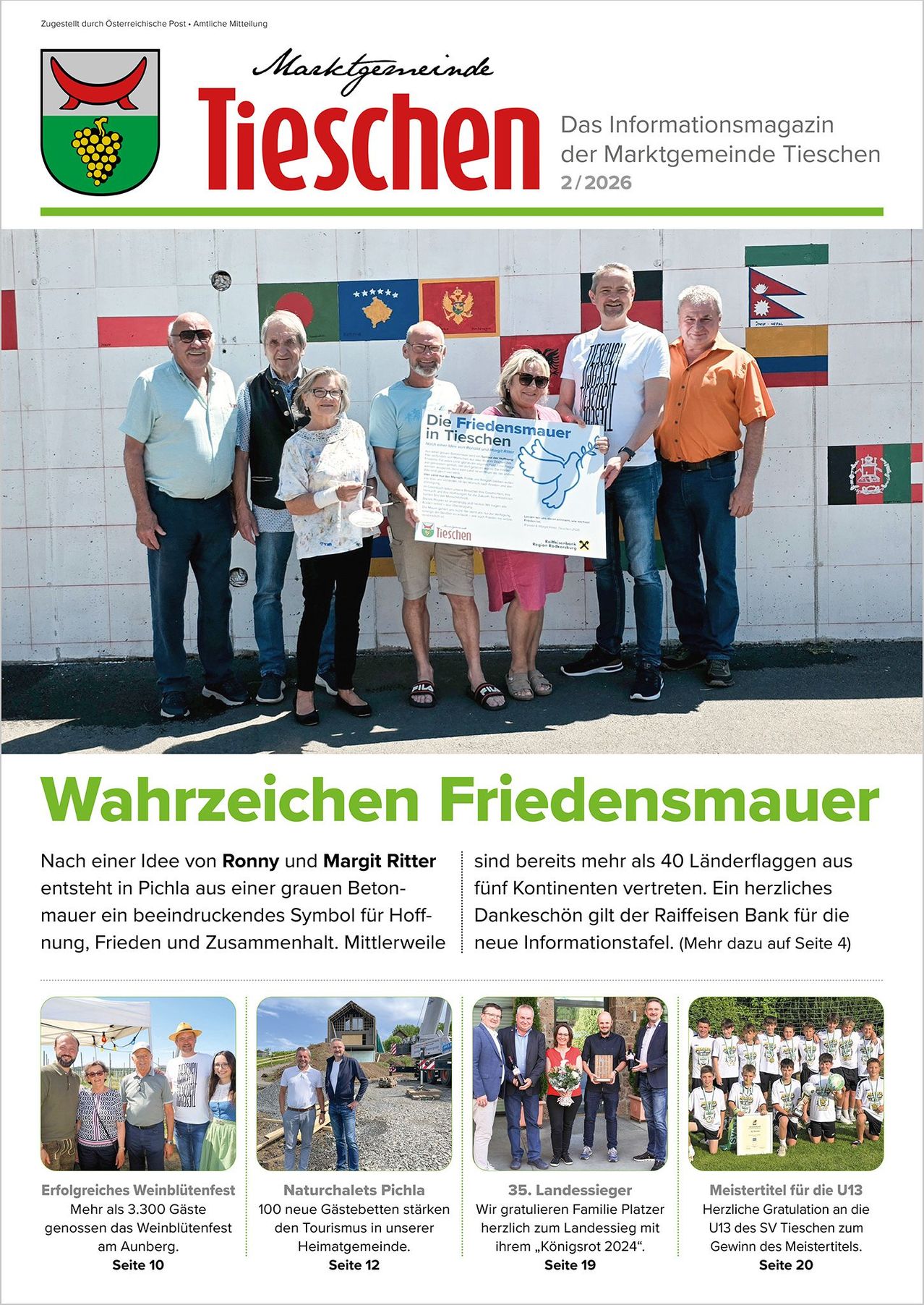 Eine Gruppe von Menschen steht vor einer Wand mit Flaggen verschiedener Länder. Sie halten ein großes Plakat mit dem Text 'Die Friedensmauer in Tischen' und einem Taubensymbol. Sie lächeln und posieren für ein Foto.