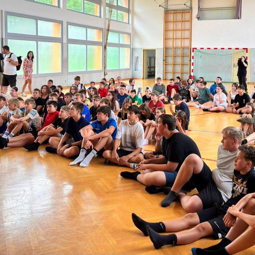 Eine Gruppe von Schülern sitzt auf dem Boden in einer Turnhalle und hört einem Sprecher zu. Einige tragen Turnschuhe und Socken.