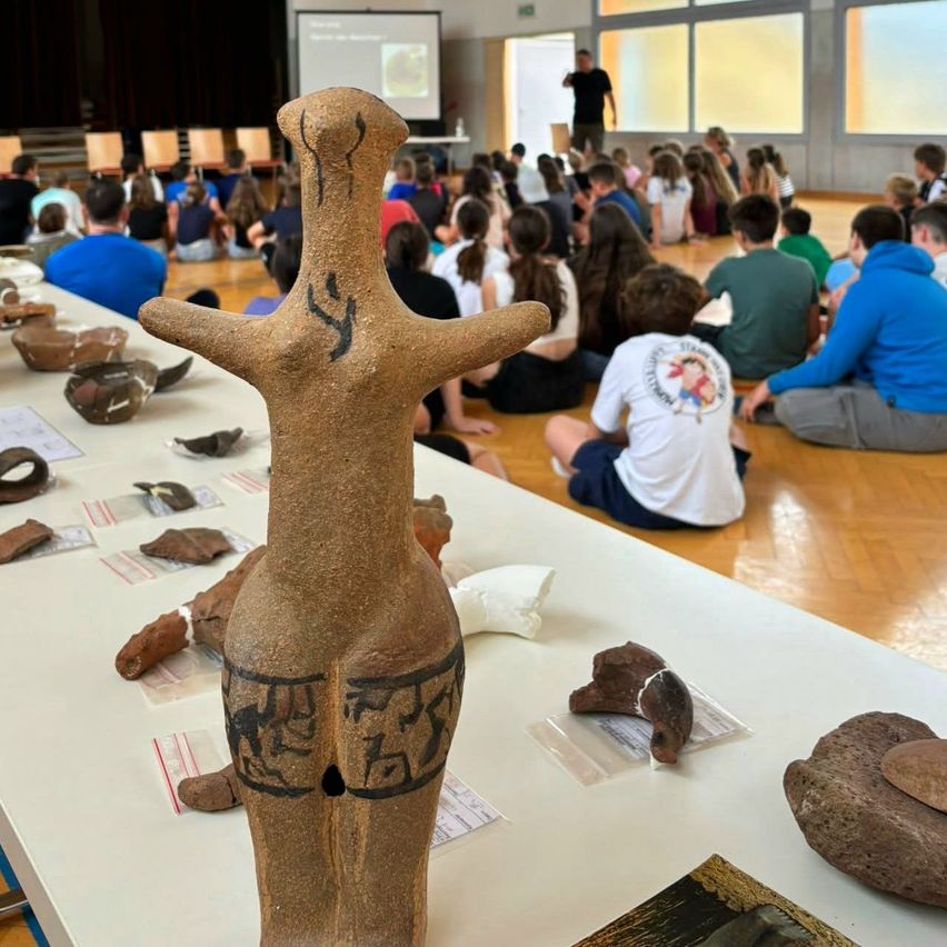 Ein Klassenzimmer mit einer großen Tonfigur auf einem Tisch, umgeben von verschiedenen Artefakten. Schüler sitzen auf dem Boden und hören einer Präsentation zu.