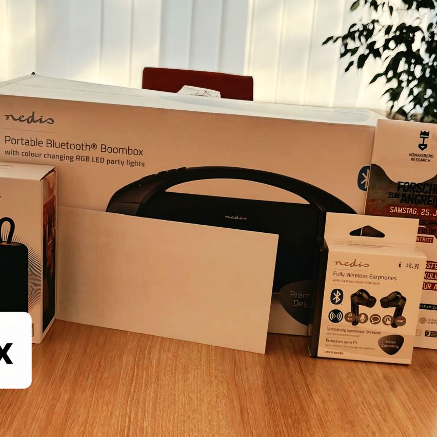 Auf einem Holztisch sind eine große Box mit der Aufschrift 'Portable Bluetooth Boombox' mit RGB LED-Lichtern sowie kleinere Boxen mit kabellosen Kopfhörern und einem Buch zu sehen.