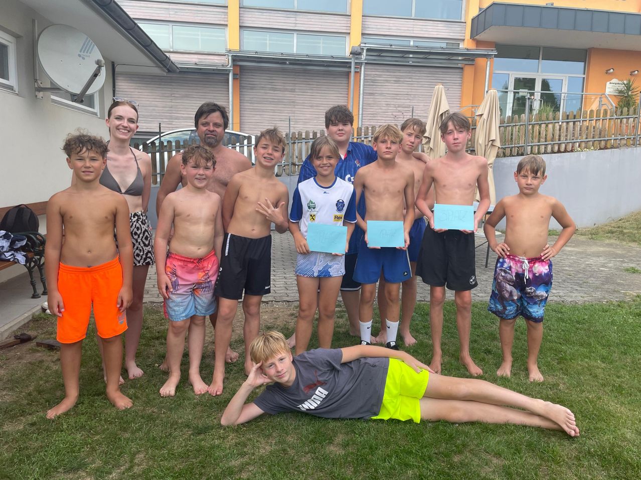 Eine Gruppe junger Jungen und einige Erwachsene stehen und sitzen auf einem Rasen, halten blaue Papiere und posieren für ein Foto. Dahinter befindet sich ein Gebäude mit Glasfenstern und einem Zaun.