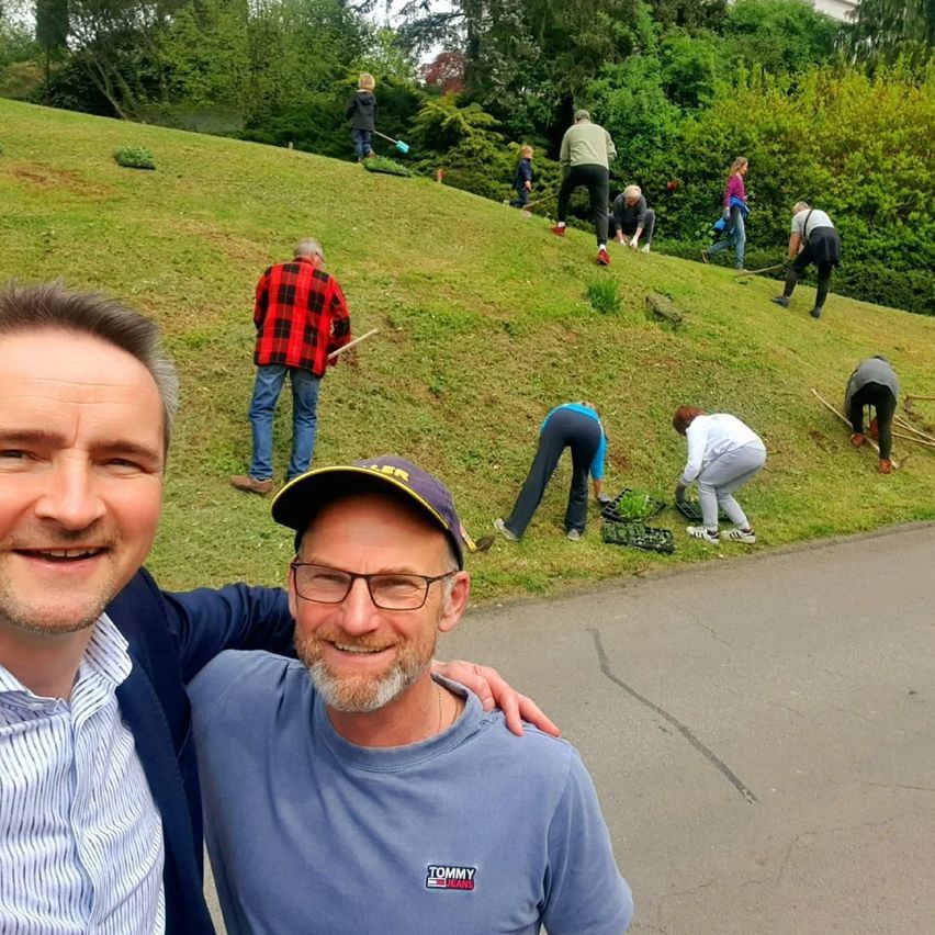 Zwei Männer lächeln für ein Foto auf einem Hügel, mit mehreren Menschen, die im Hintergrund im Garten arbeiten.