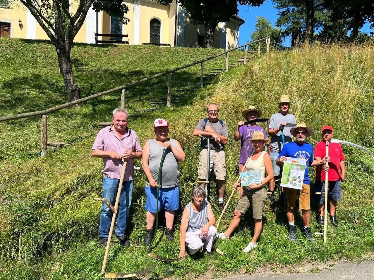 Eine Gruppe von Menschen steht in einem Grasbereich und hält Gartenwerkzeuge. Sie lächeln und posieren für ein Foto. Dahinter befindet sich ein Gebäude mit einer Treppe und einem Holzzaun.