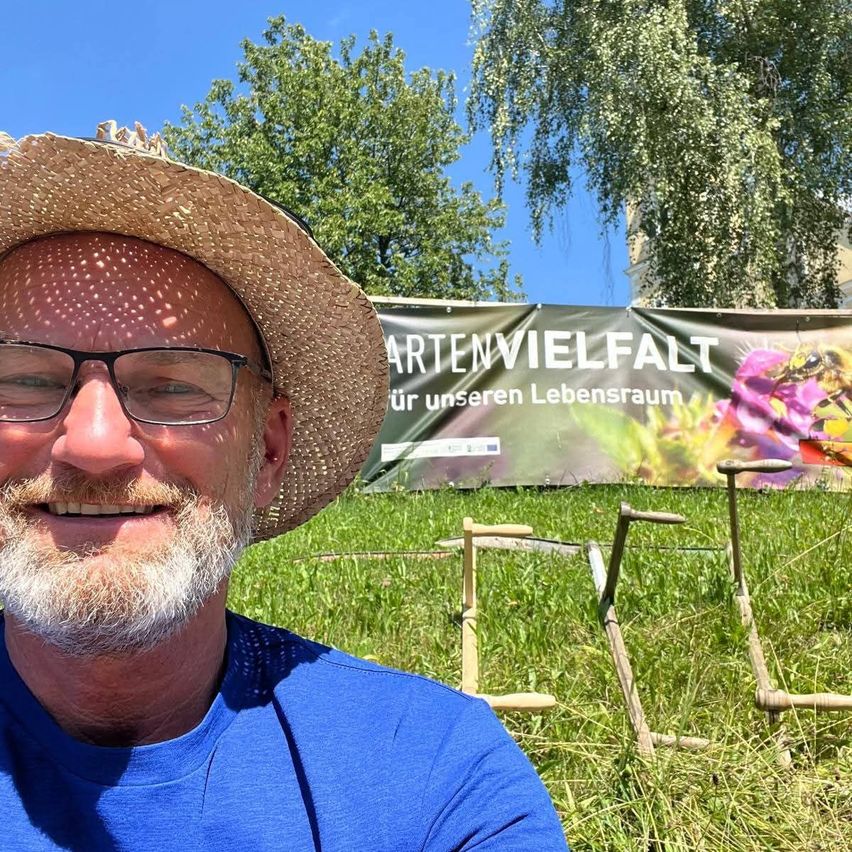 Ein Mann in einem Strohhut und mit Brille lächelt vor einem Banner, das die Artenvielfalt in einem Garten fördert.