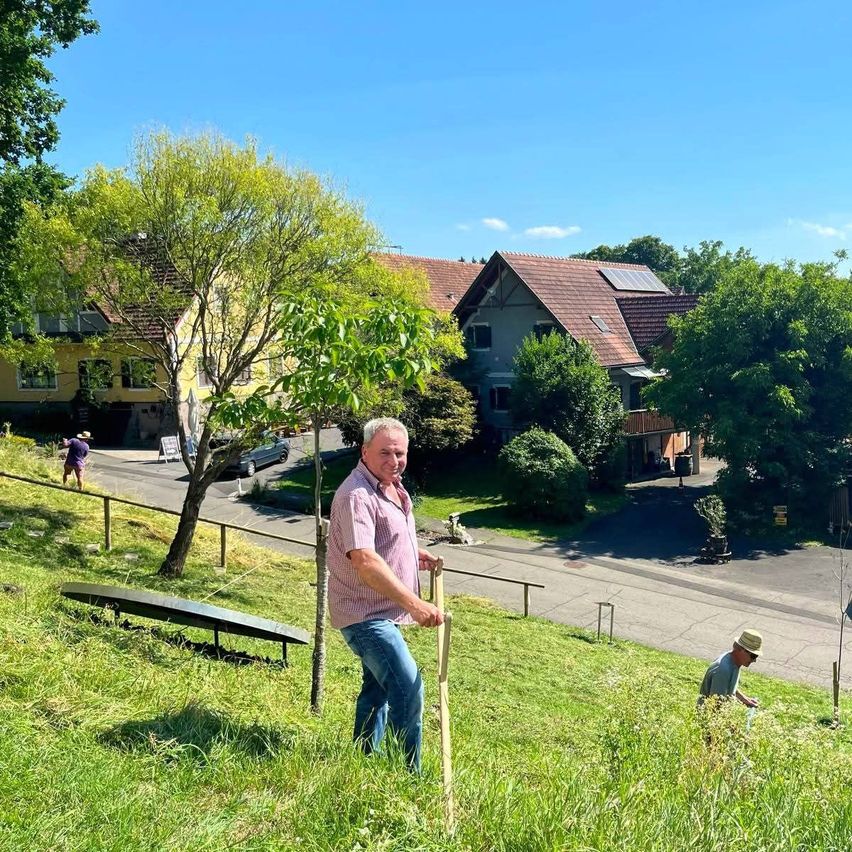 Ein Mann steht auf einem grasbewachsenen Hügel, hält einen Holzstab in der Hand und schaut in die Kamera. Hinter ihm pflanzt ein anderer Mann einen Baum. Im Hintergrund befinden sich Häuser, ein Auto und ein Baum. Der Himmel ist blau und klar.