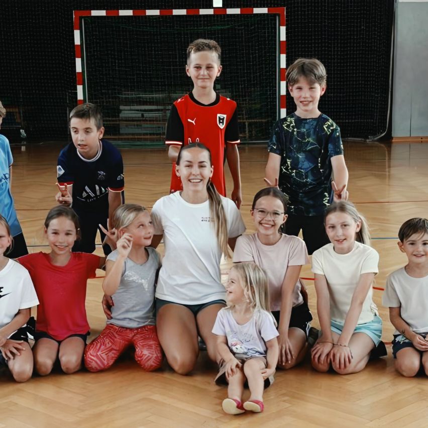 Eine Gruppe von Kindern und Jugendlichen posiert für ein Foto in einer Turnhalle. Sie tragen verschiedene Sporttrikots und Freizeitkleidung. Die Kinder lächeln und scheinen Spaß zu haben.