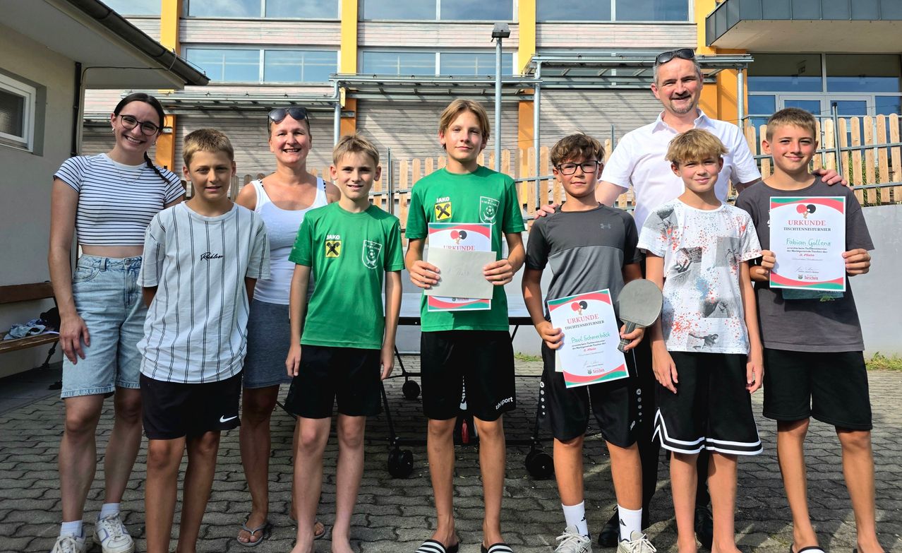 Eine Gruppe junger Jungen und Mädchen, zusammen mit zwei Erwachsenen, stehen lächelnd für ein Foto. Sie halten Urkunden und Schläger. Der Junge in der Mitte hält eine Urkunde mit einem Schläger. Hinter ihnen steht ein Gebäude mit Glasfenstern.