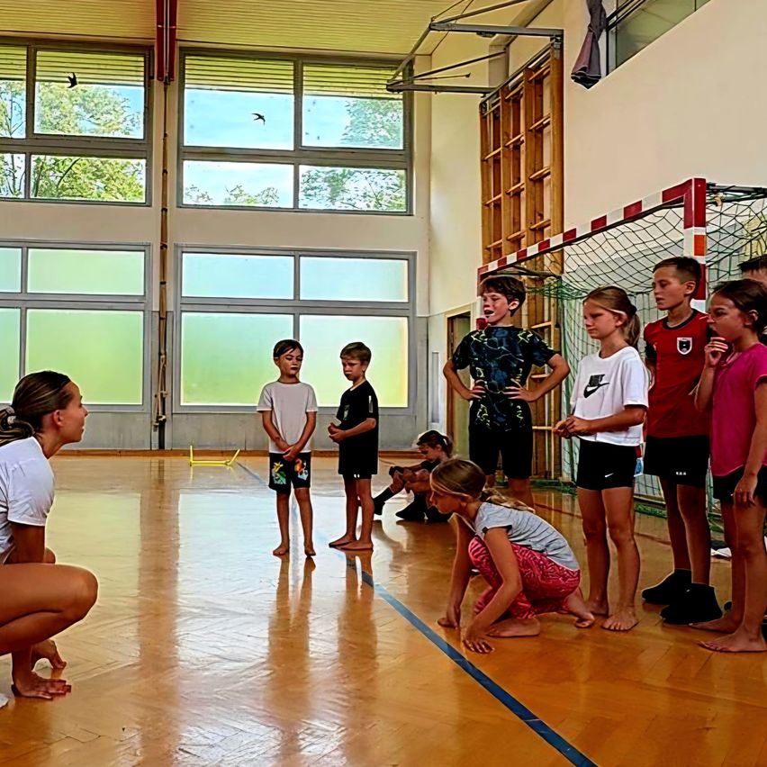 Eine Gruppe von Kindern und einer Frau versammelt sich in einer Turnhalle. Die Kinder stehen in einer Reihe, während die Frau in die Hocke geht. Sie alle schauen auf etwas vor sich. Die Turnhalle hat einen Holzboden und Glasfenster.