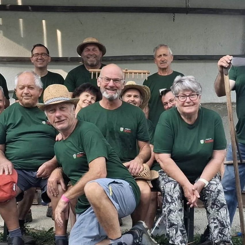 Eine Gruppe älterer Erwachsener in grünen Shirts und Hüten lächelt und posiert für ein Foto. Einige sitzen auf einer Bank, während andere stehen. Der Mann vorne kniet und lächelt.
