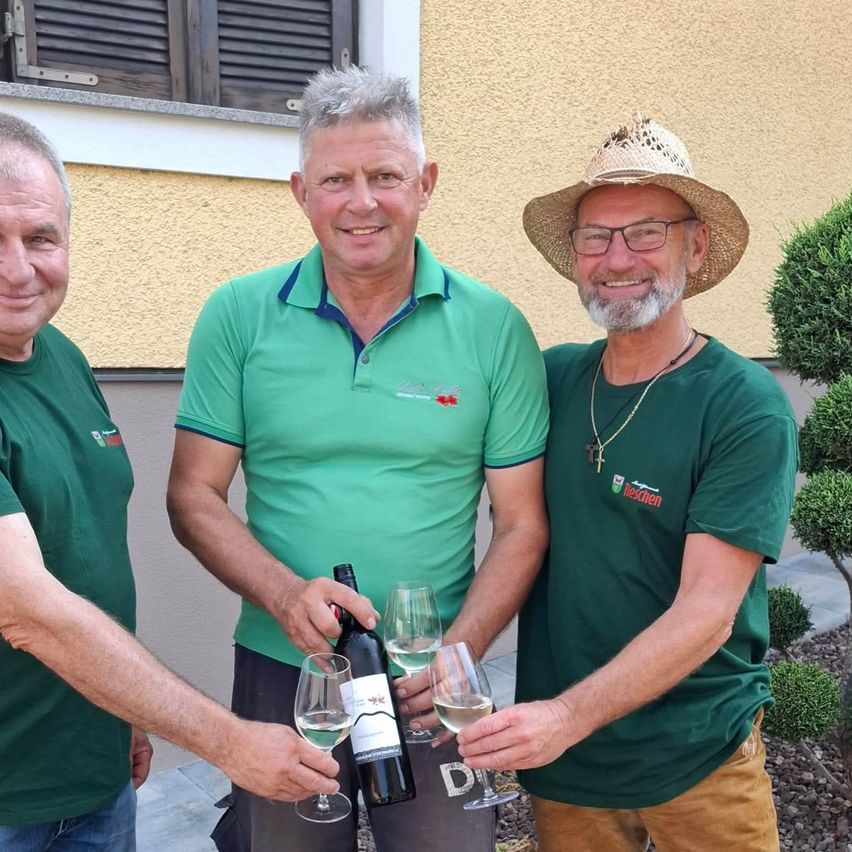 Drei Männer in grünen Shirts, einer hält eine Flasche Wein und zwei halten Weingläser, stehen vor einem Gebäude.