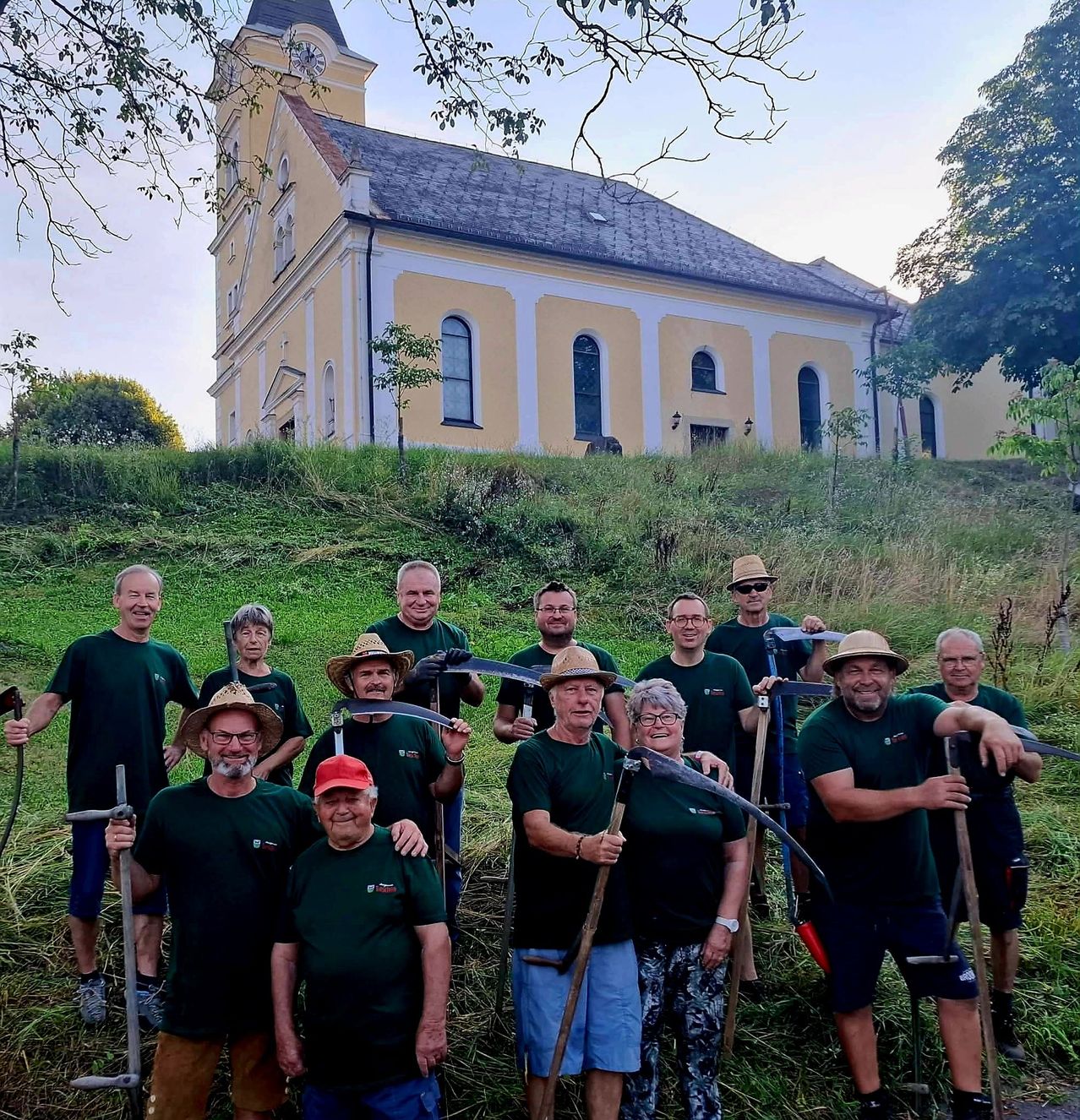Eine Gruppe von Menschen in grünen Shirts und Hüten posiert mit Sensen vor einer gelben Kirche auf einem Hügel.