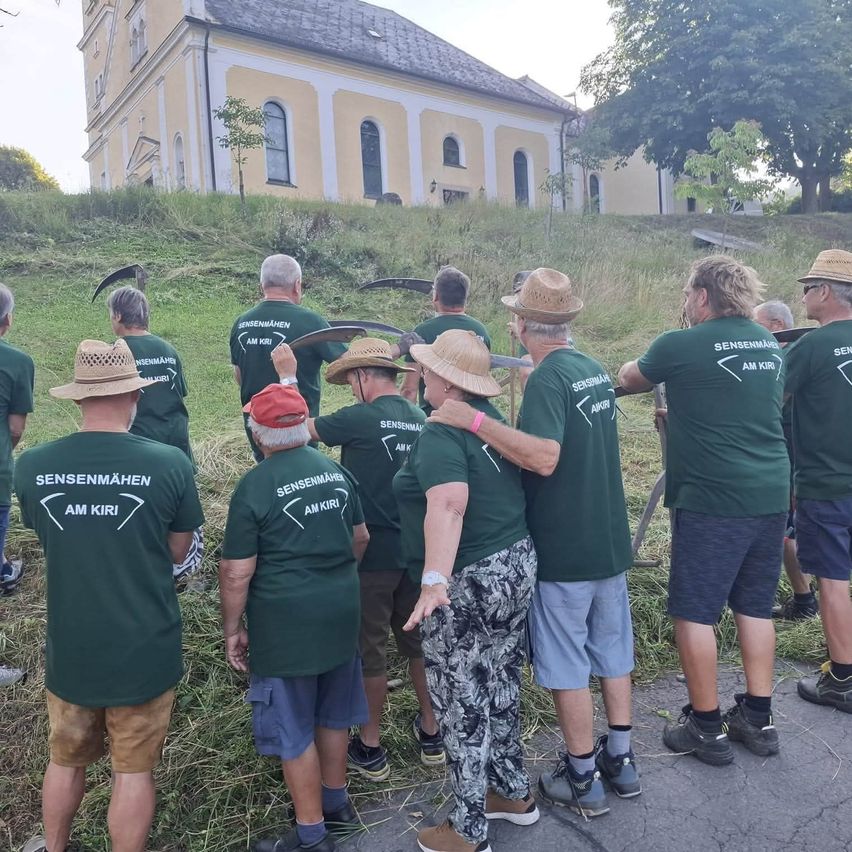 Eine Gruppe von Menschen in grünen T-Shirts mit 'Sensenmahen am Kiri' bedruckt steht auf einem Feld, möglicherweise in Vorbereitung auf eine Gemeinschaftsveranstaltung. Hinter ihnen ist eine Kirche mit einem Baum zu sehen.