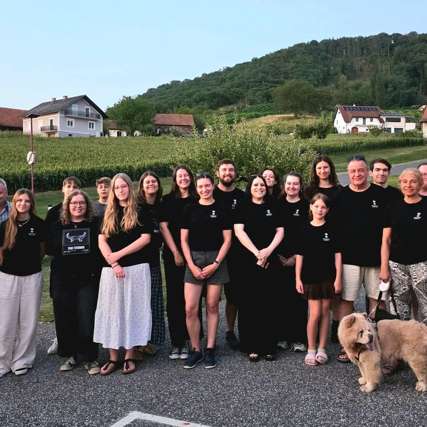 Eine Gruppe von Menschen in gleichen schwarzen T-Shirts posiert für ein Foto in einer ländlichen Umgebung mit Häusern und einem Maisfeld im Hintergrund.