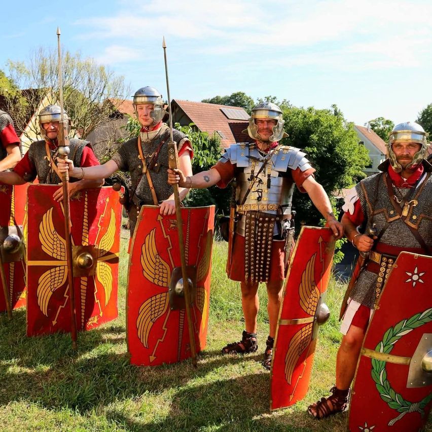 Eine Gruppe von Männern, die als römische Soldaten gekleidet sind, posiert für ein Foto, hält Schilde und Speere und hat Häuser und Bäume im Hintergrund.