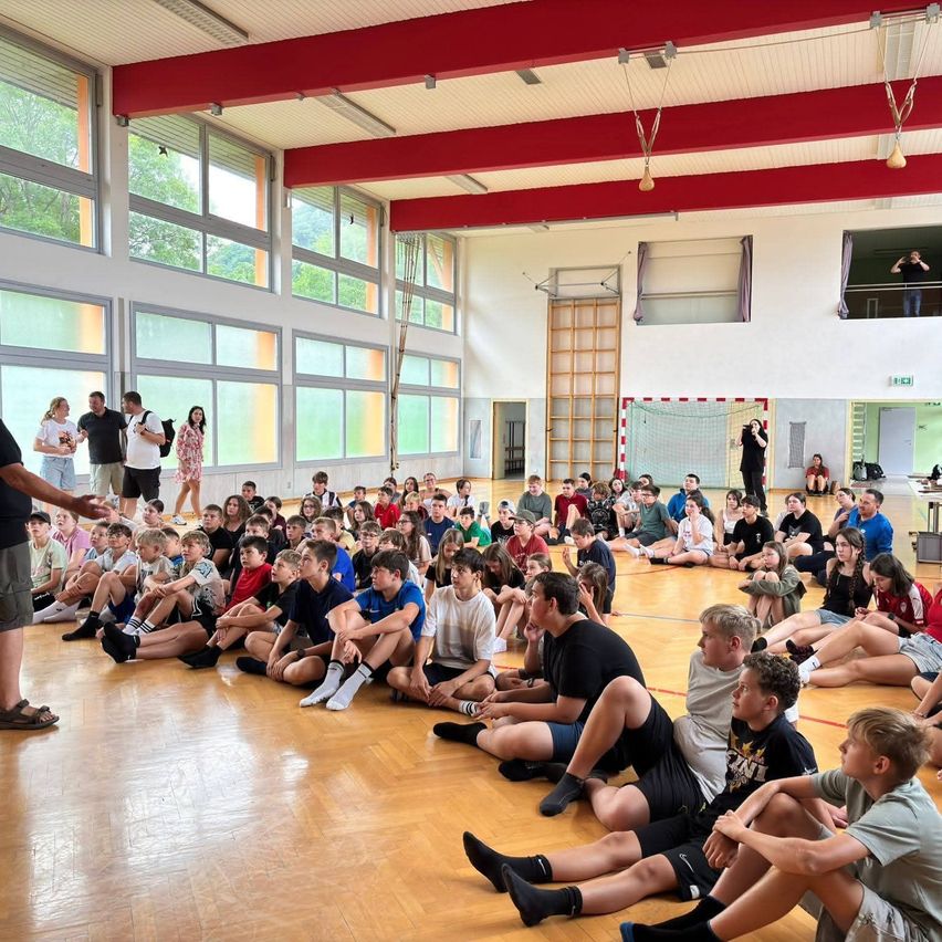 Eine große Gruppe von Menschen sitzt auf dem Boden einer Turnhalle, einige stehen herum. Ein Mann in einem schwarzen Shirt spricht die Menge an.