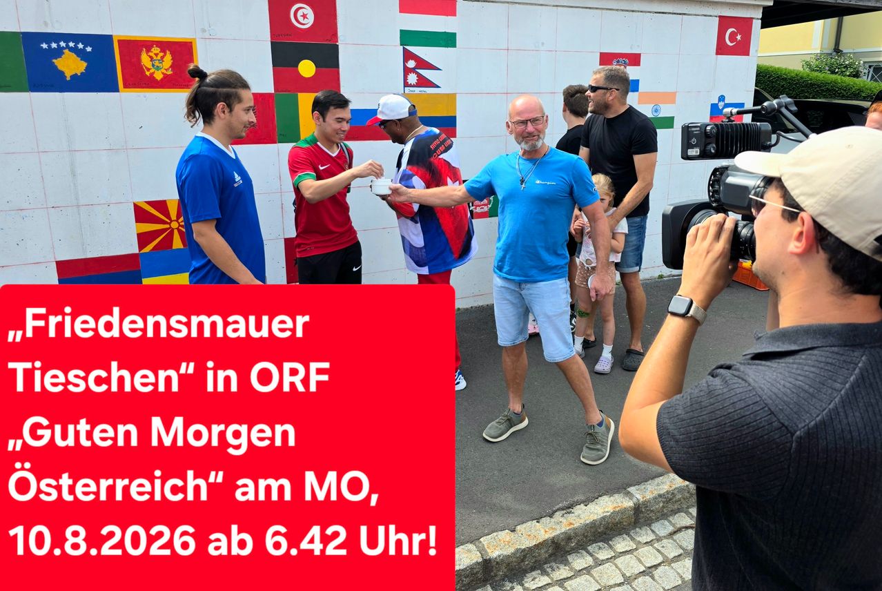 Eine Gruppe von Männern steht vor einer Wand mit Flaggen, ein Mann reicht einem anderen einen Becher, ein Mann filmt sie mit einer Kamera.