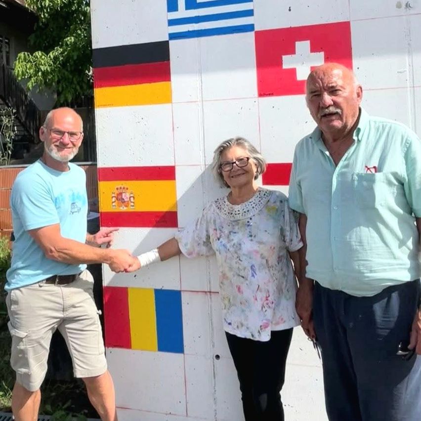 Drei Menschen stehen vor einer Wand, die mit Flaggen verschiedener Länder verziert ist. Ein Mann auf der linken Seite schüttelt die Hand einer Frau in der Mitte. Die Frau trägt eine Brille und ein Blumenhemd. Der Mann auf der rechten Seite trägt ein hellblaues Hemd.