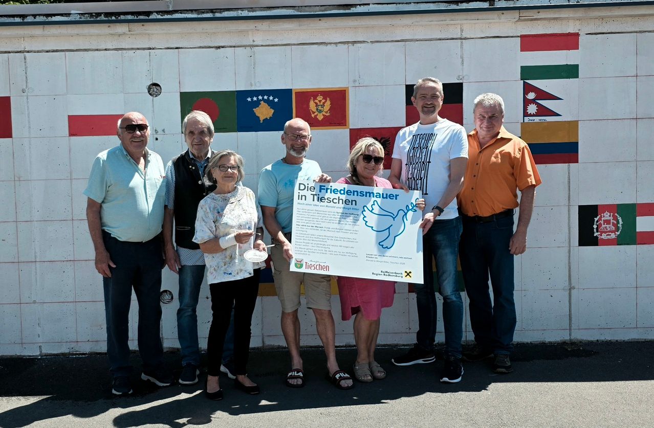 Gruppe von Erwachsenen posiert mit einem Schild mit dem Titel 'Die Friedensmauer in Tiessen' mit Flaggen hinter ihnen.