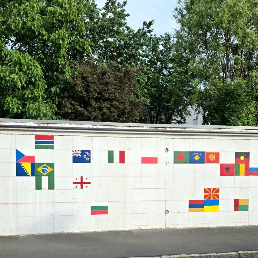 Eine weiße Wand zeigt mehrere Flaggen in einem Rastermuster, darunter Brasilien, Australien, Italien und mehr. Bäume sind im Hintergrund zu sehen.