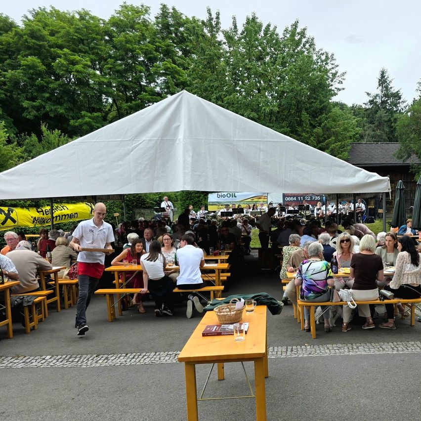 Ein großes weißes Zelt mit Menschen, die an Tischen und Bänken sitzen. Ein Kellner geht mit einem Tablett vorbei. Bäume und ein Gebäude im Hintergrund.