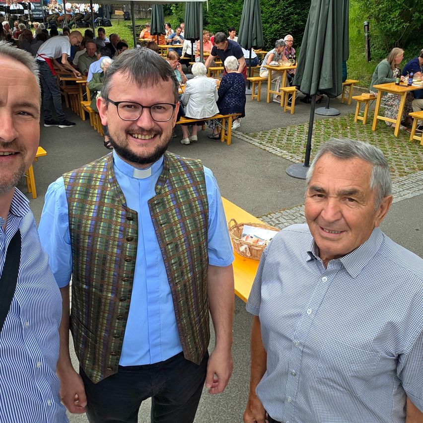 Drei Männer stehen lächelnd für ein Foto. Einer trägt eine Weste, einer ein Priesterkragen und der dritte ein blaues Hemd. Hinter ihnen ist ein Outdoor-Essbereich mit Tischen und Stühlen, an denen Menschen sitzen und stehen.