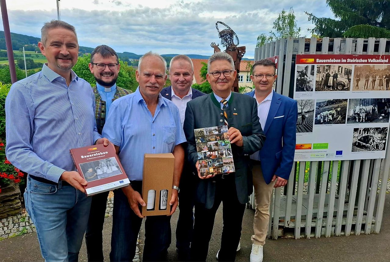 Sechs Männer stehen vor einem weißen Zaun, halten Bücher und eine Box in der Hand. Sie lächeln und schauen zur Kamera. Dahinter befindet sich ein Plakat mit Bildern.