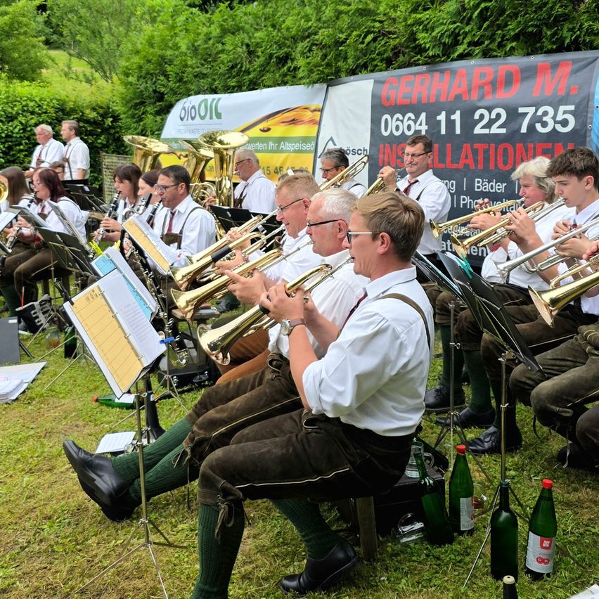 Ein Blasorchester spielt auf einem Rasenfeld mit Notenständern. Sie tragen traditionelle Kleidung und spielen Trompeten und Posaunen.