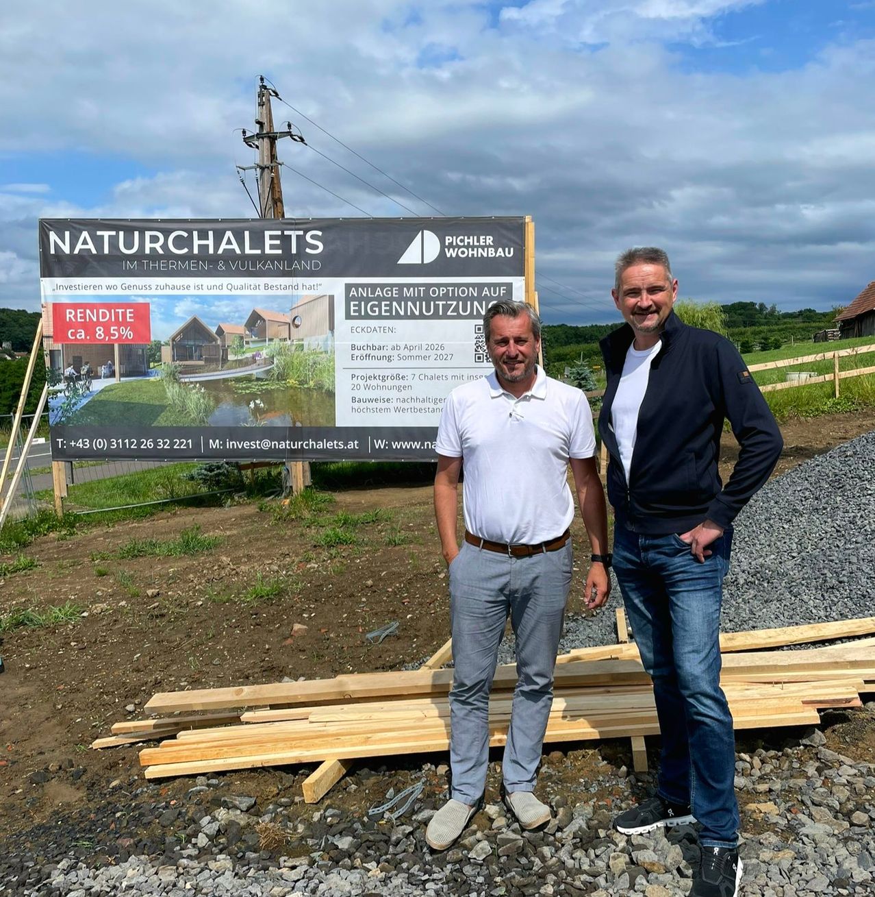 Zwei Männer stehen neben einem großen Werbeschild für Natur Chalets im Thermen- & Vulkanland. Das Schild bietet eine Rendite von 8,5% und nennt den Eröffnungstermin vom 26. April 2026.