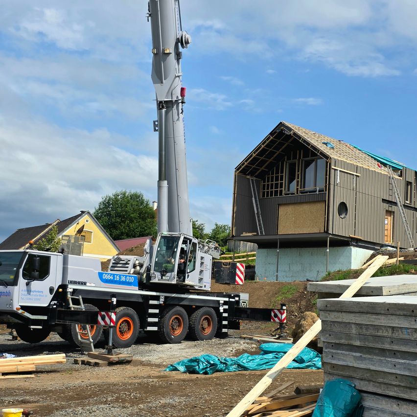 Ein Liebherr-Kran steht auf einer Baustelle, hinter ihm befindet sich ein Haus im Bau. Das Haus hat eine Leiter, ein Fenster und eine kreisförmige Öffnung. Der Boden ist mit Erde bedeckt.