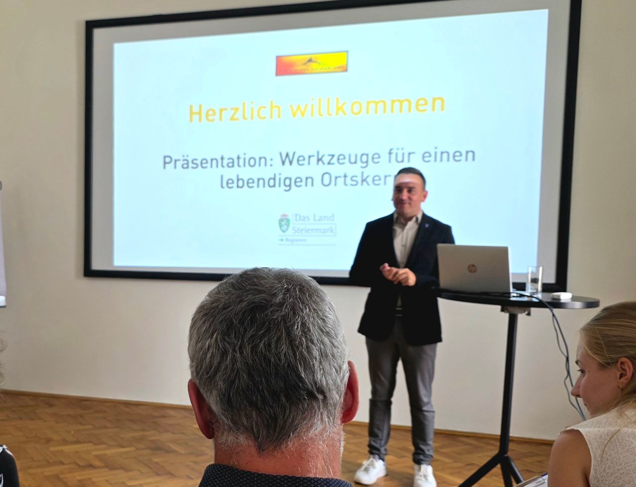 Ein Mann hält eine Präsentation über Werkzeuge für eine lebendige Ortsgemeinschaft, vor einer Projektionsleinwand stehend. Ein anderer Person sitzt davor.
