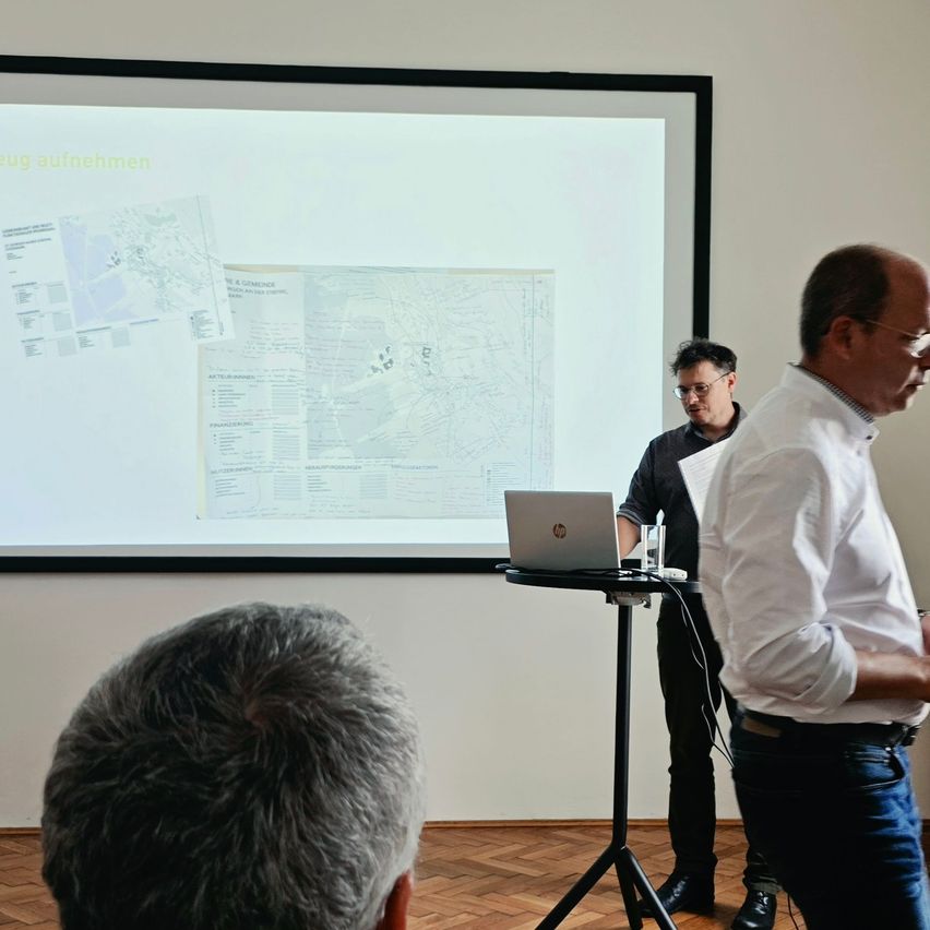 Zwei Männer stehen vor einer Projektionsleinwand mit einer Präsentation. Einer hält ein Papier, der andere einen Laptop. Eine Person schaut von hinten zu.
