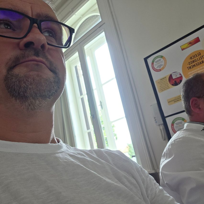 Ein Mann mit Brille schaut nach oben, wahrscheinlich in einem Büro. Hinter ihm schaut ein anderer Mann auf etwas. Ein Fenster und ein Schild mit Text sind im Hintergrund.