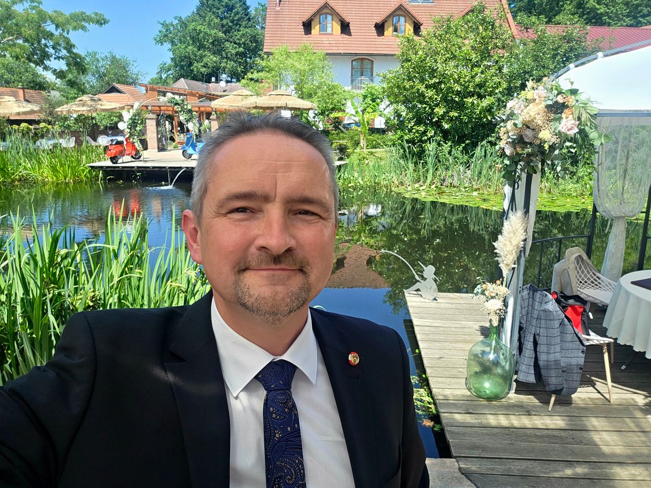 Ein Mann im Anzug steht am Teich mit einem Haus und Bäumen im Hintergrund. Er lächelt in die Kamera.
