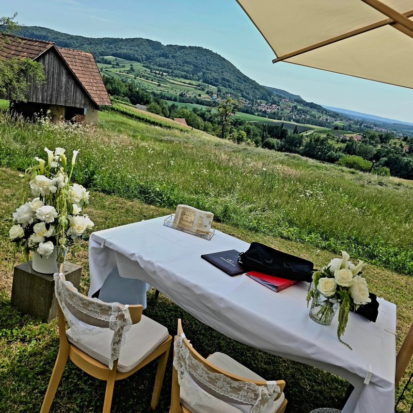 Ein weißer Tisch mit weißen Blumen auf einem Grashügel. Ein Haus und ein Berg sind im Hintergrund. Ein weißer Stoff bedeckt den Tisch.