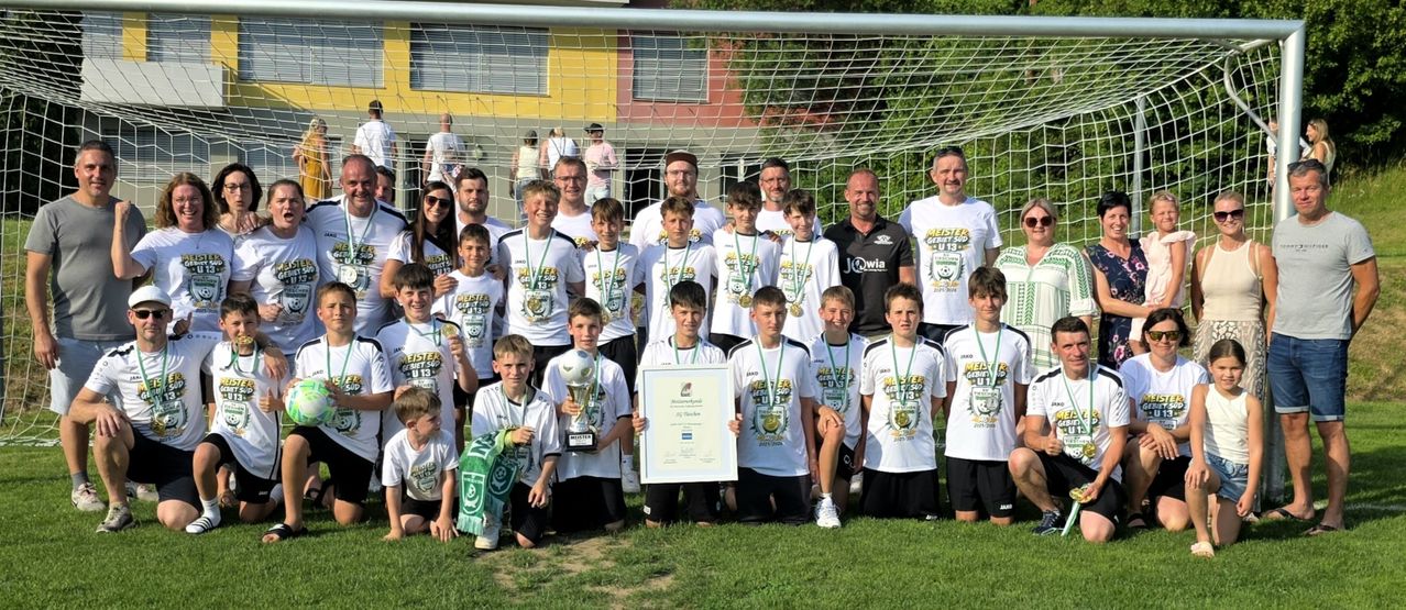Eine Fußballmannschaft posiert mit Trophäen und Medaillen vor einem Netz. Sie tragen weiße Trikots mit verschiedenen Texten und Zahlen. Zwei Männer halten ein großes Zertifikat.