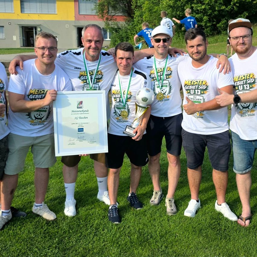 Eine Gruppe von Männern steht auf einem Feld, hält eine Trophäe und Urkunden. Sie tragen passende weiße T-Shirts und Shorts. Einige tragen eine Brille und Turnschuhe. Ein Fußball wird von einem von ihnen gehalten. Hinter ihnen steht ein Gebäude mit Fenstern und Pflanzen.